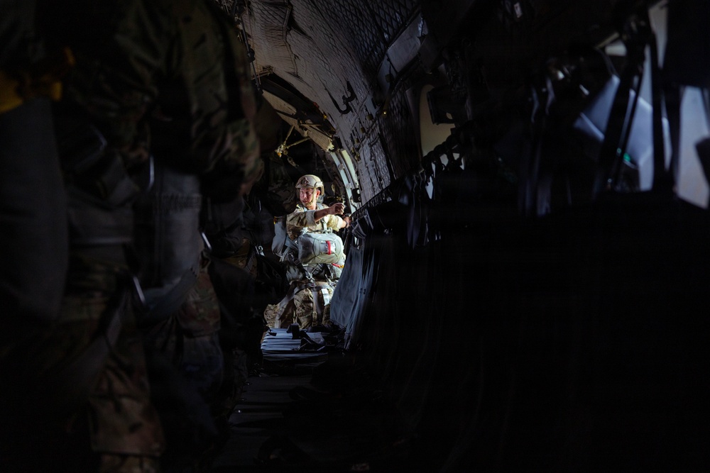 The image shows the interior of a military transport aircraft with personnel in full combat gear. A central figure, illuminated by natural or ambient light from the rear of the aircraft, appears to be a jumpmaster or aircrew member gesturing toward other soldiers seated along the fuselage walls. The scene suggests a pre-jump or in-flight briefing scenario during an airborne operation.