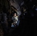 The image shows the interior of a military transport aircraft with personnel in full combat gear. A central figure, illuminated by natural or ambient light from the rear of the aircraft, appears to be a jumpmaster or aircrew member gesturing toward other soldiers seated along the fuselage walls. The scene suggests a pre-jump or in-flight briefing scenario during an airborne operation.