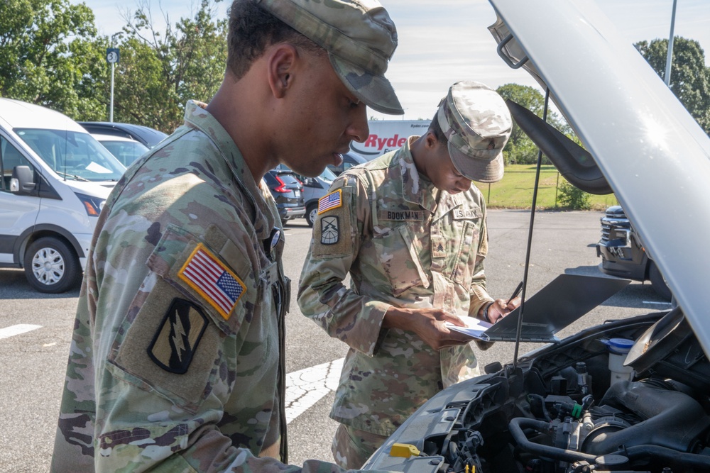 Two U.S. Army soldiers in Operational Camouflage Pattern (OCP) uniforms are conducting a vehicle inspection in a parking lot. One soldier is examining the engine compartment while the other takes notes on a small notepad or form. A Ryder truck and other vehicles are visible in the background.