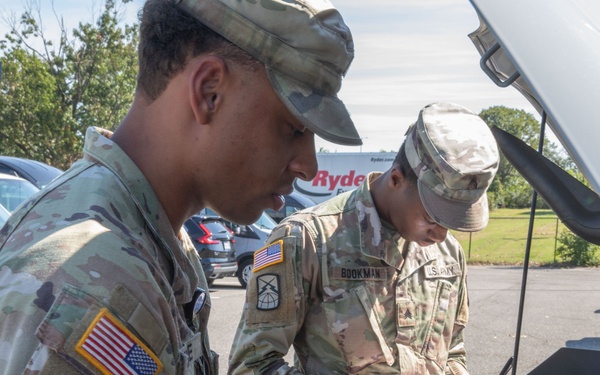 Two U.S. Army soldiers in Operational Camouflage Pattern (OCP) uniforms are conducting a vehicle inspection in a parking lot. One soldier is examining the engine compartment while the other takes notes on a small notepad or form. A Ryder truck and other vehicles are visible in the background.