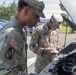 Two U.S. Army soldiers in Operational Camouflage Pattern (OCP) uniforms are conducting a vehicle inspection in a parking lot. One soldier is examining the engine compartment while the other takes notes on a small notepad or form. A Ryder truck and other vehicles are visible in the background.