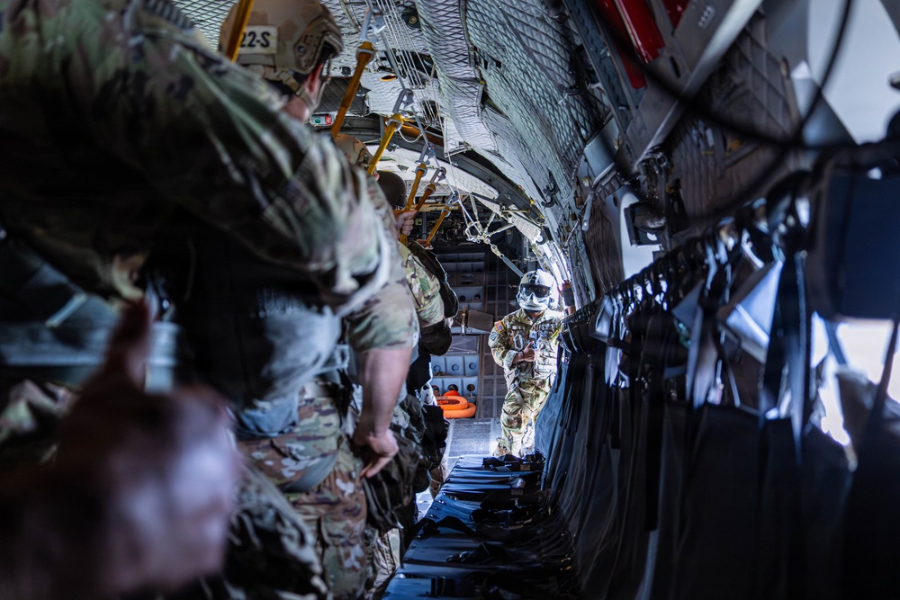 Military personnel are positioned inside what appears to be a large military transport aircraft, likely a C-130 Hercules, near an open rear cargo ramp. The soldiers are wearing full combat gear including multicam uniforms, helmets, and tactical equipment, suggesting preparation for an airborne operation or parachute jump. One individual near the open ramp is wearing a full-face helmet with visor, consistent with a jumpmaster or loadmaster role.