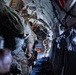 Military personnel are positioned inside what appears to be a large military transport aircraft, likely a C-130 Hercules, near an open rear cargo ramp. The soldiers are wearing full combat gear including multicam uniforms, helmets, and tactical equipment, suggesting preparation for an airborne operation or parachute jump. One individual near the open ramp is wearing a full-face helmet with visor, consistent with a jumpmaster or loadmaster role.