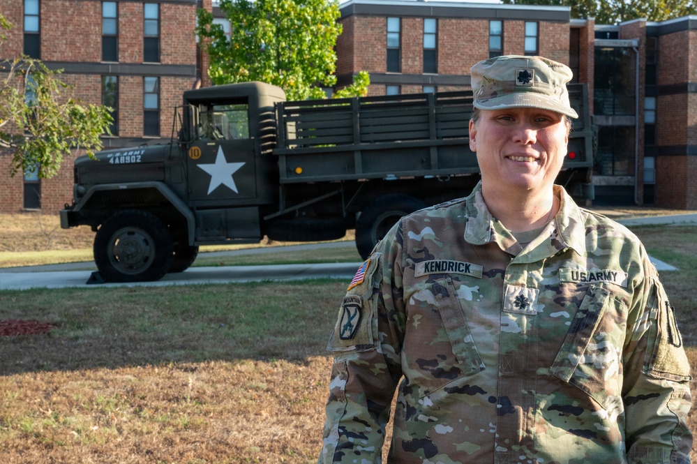 A U.S. Army soldier named Kendrick, wearing Operational Camouflage Pattern (OCP) uniform, stands in front of a vintage military truck on what appears to be a military installation. The soldier displays the rank of Lieutenant Colonel (oak leaf insignia) and wears a 10th Mountain Division patch. The setting includes brick barracks buildings in the background.