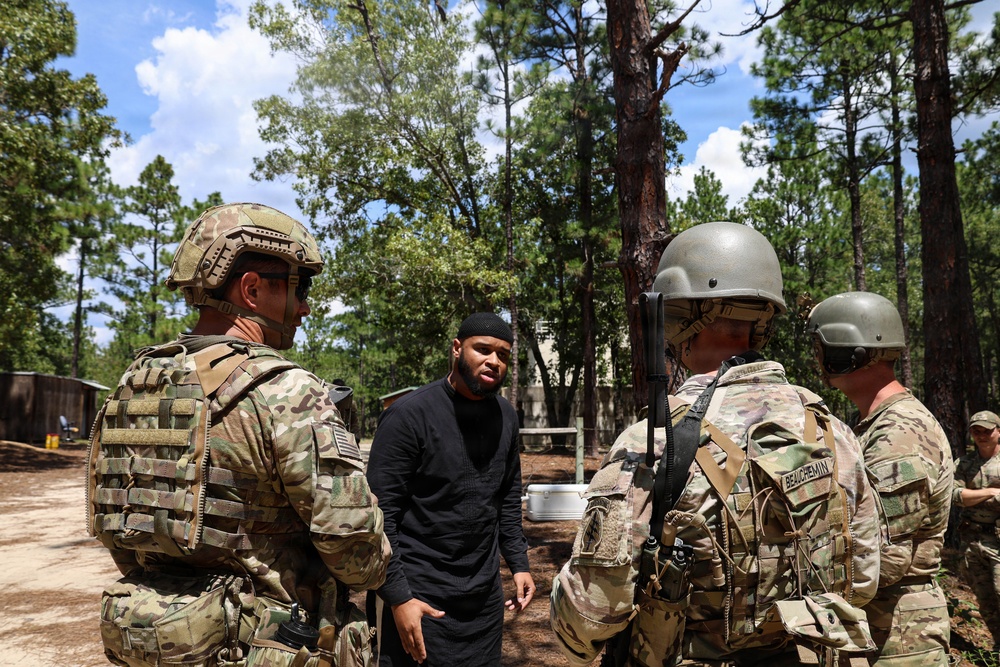 The image shows U.S. military personnel in MultiCam/OCP camouflage uniforms interacting with a civilian during what appears to be a training exercise in a wooded area. One soldier's name tape reads 'BEAUCHEMIN' on the back of their plate carrier. The setting suggests a simulated scenario or role-player interaction at a military training facility.