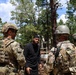 The image shows U.S. military personnel in MultiCam/OCP camouflage uniforms interacting with a civilian during what appears to be a training exercise in a wooded area. One soldier's name tape reads 'BEAUCHEMIN' on the back of their plate carrier. The setting suggests a simulated scenario or role-player interaction at a military training facility.