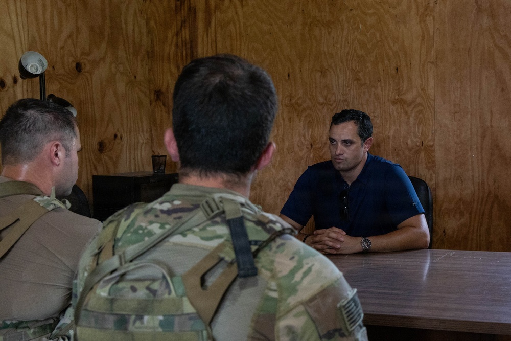 A meeting is taking place inside a room with plywood walls between a civilian and two military personnel in camouflage uniforms. The civilian, wearing a blue polo shirt, is seated across a wooden table facing the two uniformed individuals whose backs are to the camera. The setting appears to be a temporary or forward operating facility.