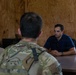 A meeting is taking place inside a room with plywood walls between a civilian and two military personnel in camouflage uniforms. The civilian, wearing a blue polo shirt, is seated across a wooden table facing the two uniformed individuals whose backs are to the camera. The setting appears to be a temporary or forward operating facility.
