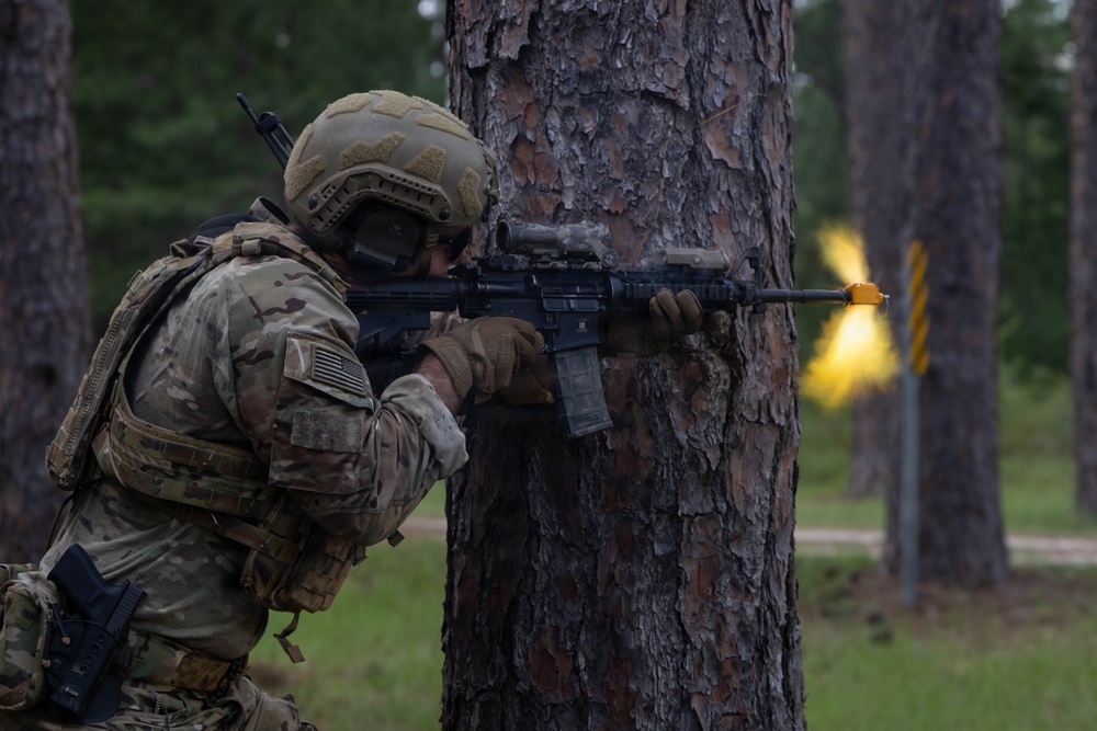A U.S. Army soldier in full combat gear is taking cover behind a large pine tree while aiming an assault rifle. A muzzle flash or bright yellow flame is visible downrange, suggesting active fire or a training exercise. The setting appears to be a wooded training area, consistent with a military exercise environment.
