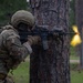 A U.S. Army soldier in full combat gear is taking cover behind a large pine tree while aiming an assault rifle. A muzzle flash or bright yellow flame is visible downrange, suggesting active fire or a training exercise. The setting appears to be a wooded training area, consistent with a military exercise environment.
