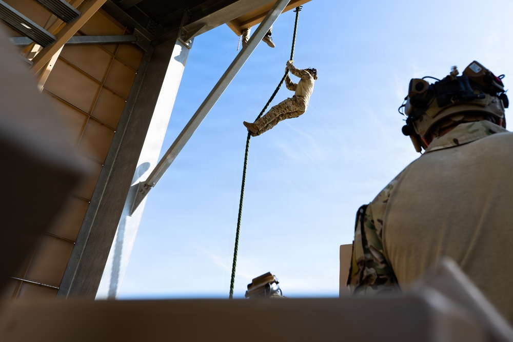 A military personnel in desert camouflage uniform is fast-roping or rappelling down a rope from an elevated structure, likely a helicopter or ship deck. A second service member in multicam/OCP uniform with a tactical helmet and communications equipment observes from the foreground. The setting appears to be a naval vessel or elevated platform with ocean visible in the background.