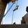TACP students receive rappel training A military personnel in desert camouflage uniform is fast-roping or rappelling down a rope from an elevated structure, likely a helicopter or ship deck. A second service member in multicam/OCP uniform with a tactical helmet and communications equipment observes from the foreground. The setting appears to be a naval vessel or elevated platform with ocean visible in the background.