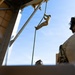 A military personnel in desert camouflage uniform is fast-roping or rappelling down a rope from an elevated structure, likely a helicopter or ship deck. A second service member in multicam/OCP uniform with a tactical helmet and communications equipment observes from the foreground. The setting appears to be a naval vessel or elevated platform with ocean visible in the background.