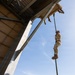 A military or law enforcement personnel in camouflage uniform is rappelling down a rope from the edge of a large building or training structure. The photo is taken from a low angle looking upward, capturing the individual mid-descent against a clear blue sky. A second person is partially visible at the top edge of the structure, likely serving as an anchor or instructor.