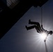 A silhouetted figure is rappelling or abseiling down the side of a large building, captured from a low angle looking upward. The sun creates a dramatic starburst effect directly behind the person, backlighting the scene against a blue sky. The image is a striking action shot emphasizing contrast between the dark structure and bright sky.