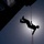 TACP students receive rappel training A silhouetted person is rappelling or abseiling down the side of a large structure, suspended by an orange rope against a bright backlit sky. The image is taken from a low angle looking upward, creating a dramatic contrast between the dark silhouette and the glowing sun behind clouds. The structure visible in the upper left appears to be a tall building or bridge.