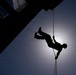 A silhouetted person is rappelling or abseiling down the side of a large structure, suspended by an orange rope against a bright backlit sky. The image is taken from a low angle looking upward, creating a dramatic contrast between the dark silhouette and the glowing sun behind clouds. The structure visible in the upper left appears to be a tall building or bridge.
