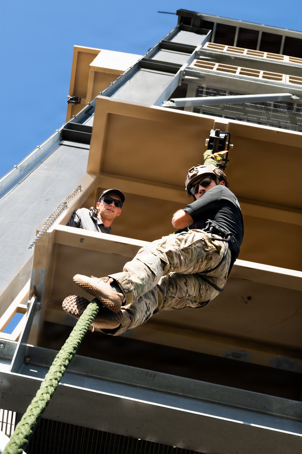 A person in military-style camouflage clothing and a helmet is rappelling down the side of a multi-story training tower using a green rope. A second person wearing a black cap and sunglasses observes from a platform above. The image is taken from a low angle looking upward, emphasizing the height and the rappelling action.