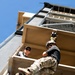 A person in military-style camouflage clothing and a helmet is rappelling down the side of a multi-story training tower using a green rope. A second person wearing a black cap and sunglasses observes from a platform above. The image is taken from a low angle looking upward, emphasizing the height and the rappelling action.