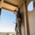 TACP students receive rappel training A person wearing military camouflage pants and a gray t-shirt stands at the edge of an elevated platform, leaning forward to look down. A rope with knots is visible in the foreground, anchored to the platform floor. The setting appears to be a military training tower with a base installation visible in the background.