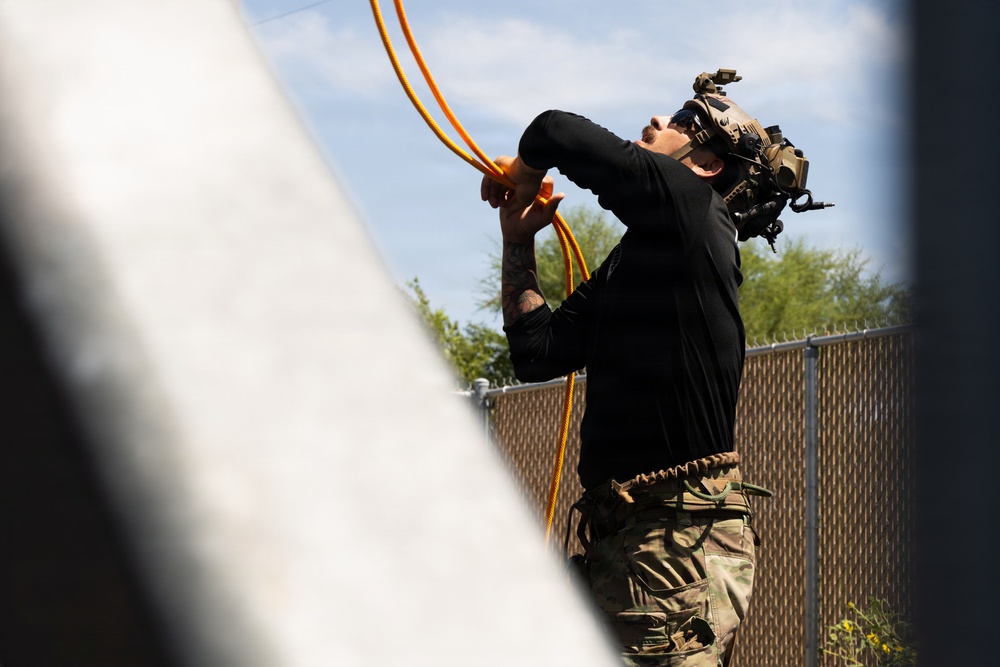 A tactical operator wearing military/law enforcement gear, including a helmet-mounted night vision device and multicam pants, is handling orange hoses or cables while looking upward. The scene appears to be an outdoor training or operational environment near a chain-link fence. The individual is dressed in a black long-sleeve shirt and camouflage pants with visible tattoos on the forearm.