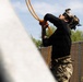 A tactical operator wearing military/law enforcement gear, including a helmet-mounted night vision device and multicam pants, is handling orange hoses or cables while looking upward. The scene appears to be an outdoor training or operational environment near a chain-link fence. The individual is dressed in a black long-sleeve shirt and camouflage pants with visible tattoos on the forearm.