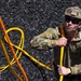 A military personnel wearing multicam camouflage uniform and tactical helmet is handling bright yellow/orange ropes or cords on a surface covered with black rubber mulch or shredded material. The individual is equipped with tactical gear including sunglasses, a combat helmet with chin strap, and appears to be managing or coiling the rope. The image is taken from a slightly elevated angle in bright daylight conditions.