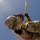 TACP students receive rappel training A military personnel in full camouflage uniform is performing a rappelling or rope descent operation against a blue sky background. The soldier is equipped with tactical gear including a helmet, protective eyewear, and black gloves, and is gripping an orange rope connected to a carabiner. The low-angle upward perspective creates a dynamic composition emphasizing the height and physical nature of the activity.