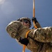 A military personnel in full camouflage uniform is performing a rappelling or rope descent operation against a blue sky background. The soldier is equipped with tactical gear including a helmet, protective eyewear, and black gloves, and is gripping an orange rope connected to a carabiner. The low-angle upward perspective creates a dynamic composition emphasizing the height and physical nature of the activity.