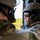 TACP students receive rappel training Two military personnel are seated inside what appears to be a helicopter in flight, looking out through an open door or window. The soldier in focus wears a tan tactical helmet, reflective ballistic goggles, communications headset, and OCP/MultiCam camouflage uniform. The goggles reflect a landscape below, consistent with aerial observation during a flight mission.