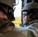 Two military personnel are seated inside what appears to be a helicopter in flight, looking out through an open door or window. The soldier in focus wears a tan tactical helmet, reflective ballistic goggles, communications headset, and OCP/MultiCam camouflage uniform. The goggles reflect a landscape below, consistent with aerial observation during a flight mission.