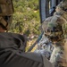 Military personnel in full combat gear are seated inside a helicopter in flight, looking out over a densely wooded, hilly terrain below. At least three individuals wearing Operational Camouflage Pattern (OCP) uniforms and tactical helmets with communication equipment are visible. One helmet bears a visible 'TACP' label, indicating Tactical Air Control Party personnel.