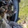 TACP students receive rappel training Military personnel in multicam/OCP camouflage uniforms are positioned at the open door of a helicopter in flight, looking down at dense green vegetation below. At least three individuals are visible wearing tactical helmets, body armor, and gloves, with one appearing to gesture or signal. The image is taken from inside the aircraft looking outward, capturing both the crew and the aerial terrain below.