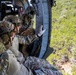 Military personnel in multicam/OCP camouflage uniforms are positioned at the open door of a helicopter in flight, looking down at dense green vegetation below. At least three individuals are visible wearing tactical helmets, body armor, and gloves, with one appearing to gesture or signal. The image is taken from inside the aircraft looking outward, capturing both the crew and the aerial terrain below.