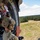 TACP students receive rappel training A military personnel in full combat gear sits at the open door of a helicopter in flight, observing the terrain below. The soldier is wearing MultiCam/OCP camouflage, a tactical helmet with hearing protection, and black gloves. The landscape visible below consists of open grassland with scattered trees and a dirt road.