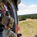 A military personnel in full combat gear sits at the open door of a helicopter in flight, observing the terrain below. The soldier is wearing MultiCam/OCP camouflage, a tactical helmet with hearing protection, and black gloves. The landscape visible below consists of open grassland with scattered trees and a dirt road.