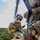 TACP students receive rappel training A military or special operations personnel is rappelling from a helicopter, suspended on a rope outside the open door of the aircraft. A second individual, visible in the foreground, appears to be a spotter or safety observer positioned at the helicopter door. The terrain below shows open grassland with tree-covered hills and what appear to be vehicles on a dirt road.