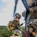 A military or special operations personnel is rappelling from a helicopter, suspended on a rope outside the open door of the aircraft. A second individual, visible in the foreground, appears to be a spotter or safety observer positioned at the helicopter door. The terrain below shows open grassland with tree-covered hills and what appear to be vehicles on a dirt road.