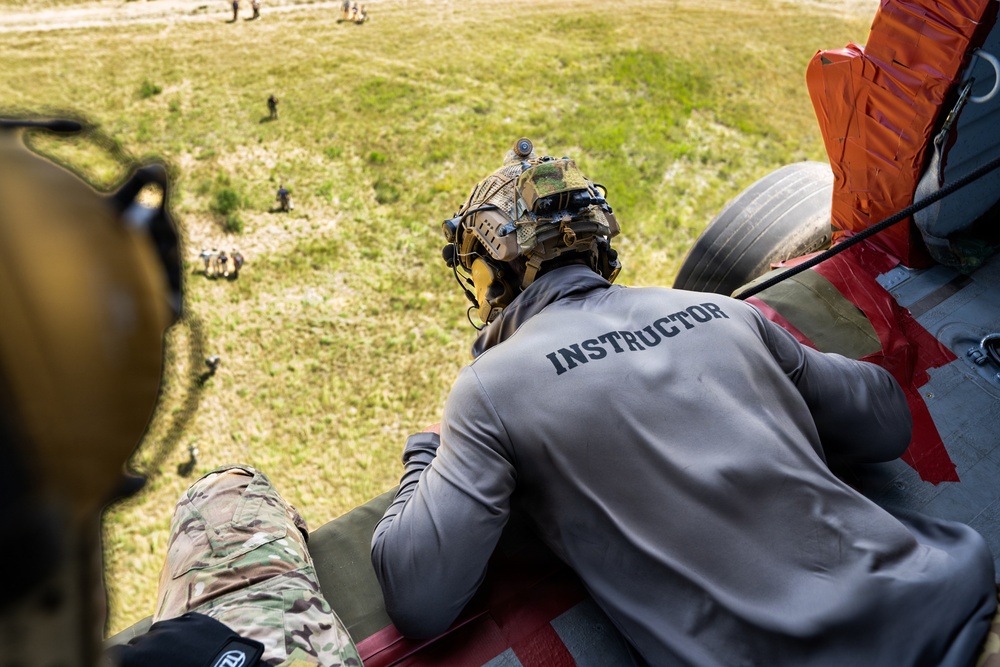 An instructor wearing a gray long-sleeve shirt labeled 'INSTRUCTOR' and a tactical helmet is positioned at the open rear ramp of a military helicopter, looking down at a grassy field below. Several personnel and equipment are visible on the ground. The image appears to document a military training exercise involving aerial operations.