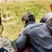 An instructor wearing a gray long-sleeve shirt labeled 'INSTRUCTOR' and a tactical helmet is positioned at the open rear ramp of a military helicopter, looking down at a grassy field below. Several personnel and equipment are visible on the ground. The image appears to document a military training exercise involving aerial operations.