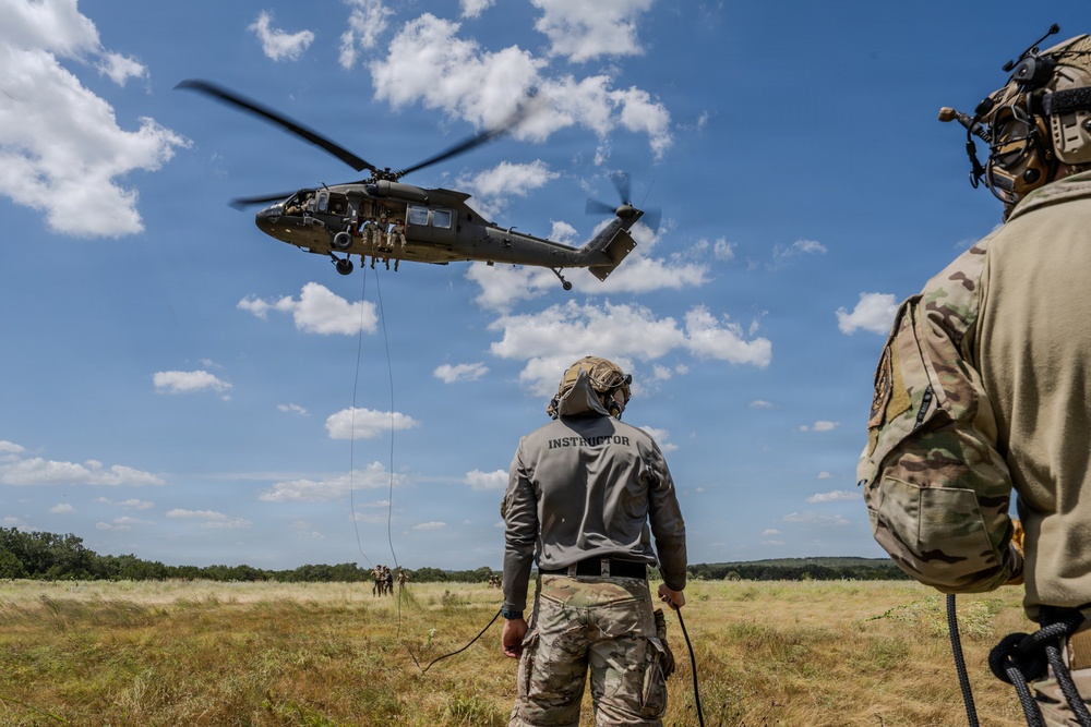 A military training exercise is underway in an open field, featuring a UH-60 Black Hawk helicopter hovering at low altitude with personnel visible in the open door. Two ground personnel are observing the operation, one clearly labeled 'INSTRUCTOR' on their jacket, while ropes hang from the helicopter suggesting a rappelling or fast-rope insertion/extraction (FRIES) exercise. Additional personnel can be seen in the background on the ground.