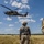 TACP students receive rappel training A military training exercise is underway in an open field, featuring a UH-60 Black Hawk helicopter hovering at low altitude with personnel visible in the open door. Two ground personnel are observing the operation, one clearly labeled 'INSTRUCTOR' on their jacket, while ropes hang from the helicopter suggesting a rappelling or fast-rope insertion/extraction (FRIES) exercise. Additional personnel can be seen in the background on the ground.