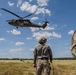 A military training exercise is underway in an open field, featuring a UH-60 Black Hawk helicopter hovering at low altitude with personnel visible in the open door. Two ground personnel are observing the operation, one clearly labeled 'INSTRUCTOR' on their jacket, while ropes hang from the helicopter suggesting a rappelling or fast-rope insertion/extraction (FRIES) exercise. Additional personnel can be seen in the background on the ground.