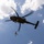 TACP students receive rappel training A military helicopter is hovering in a partly cloudy blue sky while a soldier rappels or fast-ropes down from the aircraft on a rope. The helicopter appears to be a utility/transport type with a dark olive/grey paint scheme. This appears to be a military training or operational insertion exercise.