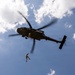 A military helicopter is hovering in a partly cloudy blue sky while a soldier rappels or fast-ropes down from the aircraft on a rope. The helicopter appears to be a utility/transport type with a dark olive/grey paint scheme. This appears to be a military training or operational insertion exercise.