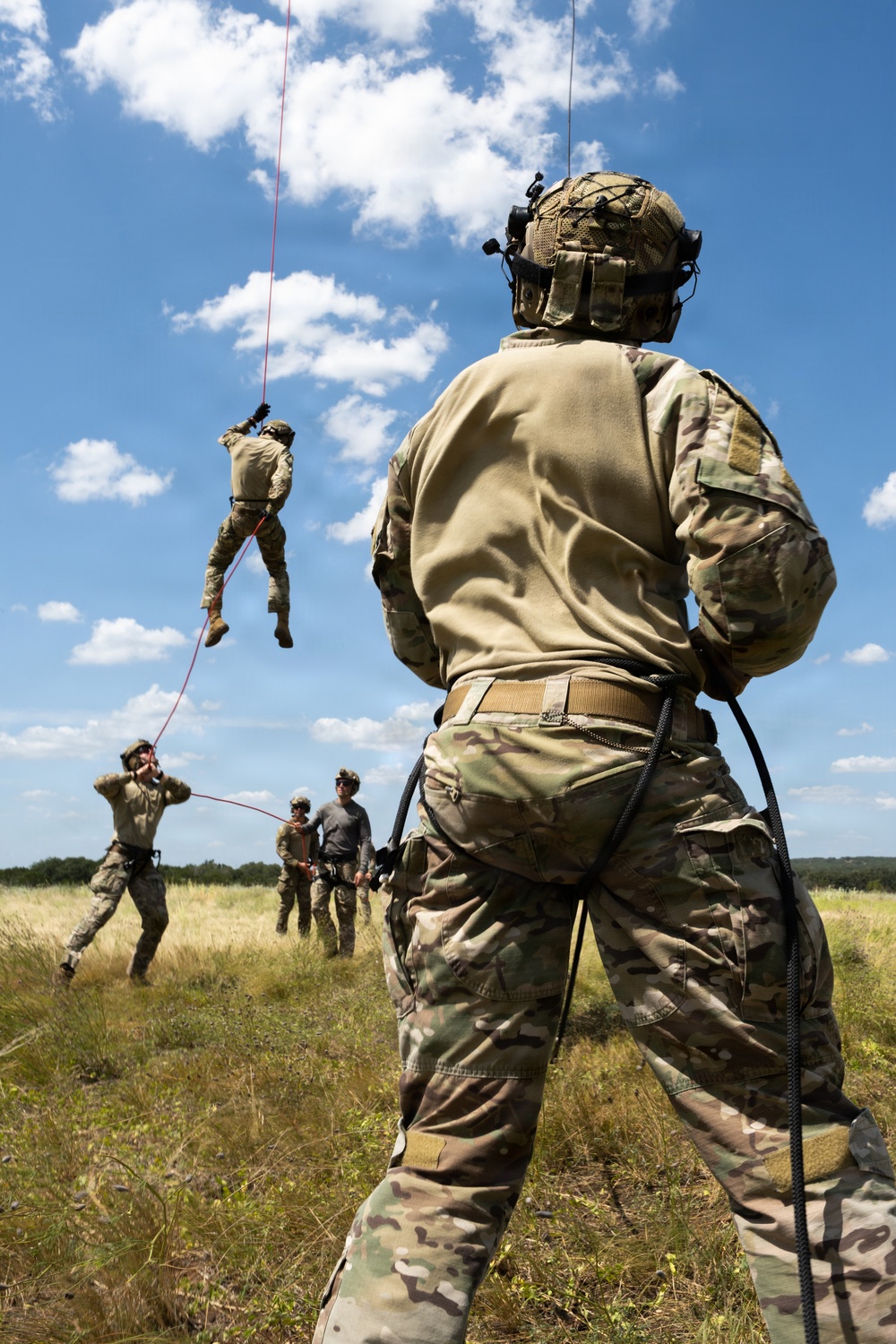 Military personnel are conducting rope/rappelling training in an open grassy field under a partly cloudy sky. One soldier is suspended mid-air on a red rope, while others on the ground observe, manage safety lines, or await their turn. A foreground soldier in full OCP (Operational Camouflage Pattern) uniform and tactical helmet is managing a belay rope.