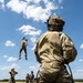 Military personnel are conducting rope/rappelling training in an open grassy field under a partly cloudy sky. One soldier is suspended mid-air on a red rope, while others on the ground observe, manage safety lines, or await their turn. A foreground soldier in full OCP (Operational Camouflage Pattern) uniform and tactical helmet is managing a belay rope.