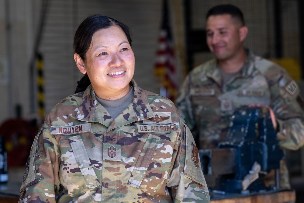 A U.S. Air Force service member named Nguyen, wearing Operational Camouflage Pattern (OCP) uniform, is smiling and looking off to the side. She displays a Chief Master Sergeant rank insignia and a flight badge on her uniform. A second service member is visible in the background, slightly out of focus, in what appears to be a hangar or maintenance facility.