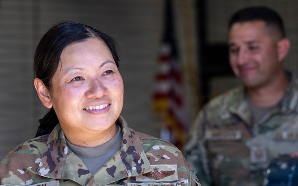 A U.S. Air Force service member named Nguyen, wearing Operational Camouflage Pattern (OCP) uniform, is smiling and looking off to the side. She displays a Chief Master Sergeant rank insignia and a flight badge on her uniform. A second service member is visible in the background, slightly out of focus, in what appears to be a hangar or maintenance facility.