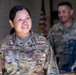 A U.S. Air Force service member named Nguyen, wearing Operational Camouflage Pattern (OCP) uniform, is smiling and looking off to the side. She displays a Chief Master Sergeant rank insignia and a flight badge on her uniform. A second service member is visible in the background, slightly out of focus, in what appears to be a hangar or maintenance facility.